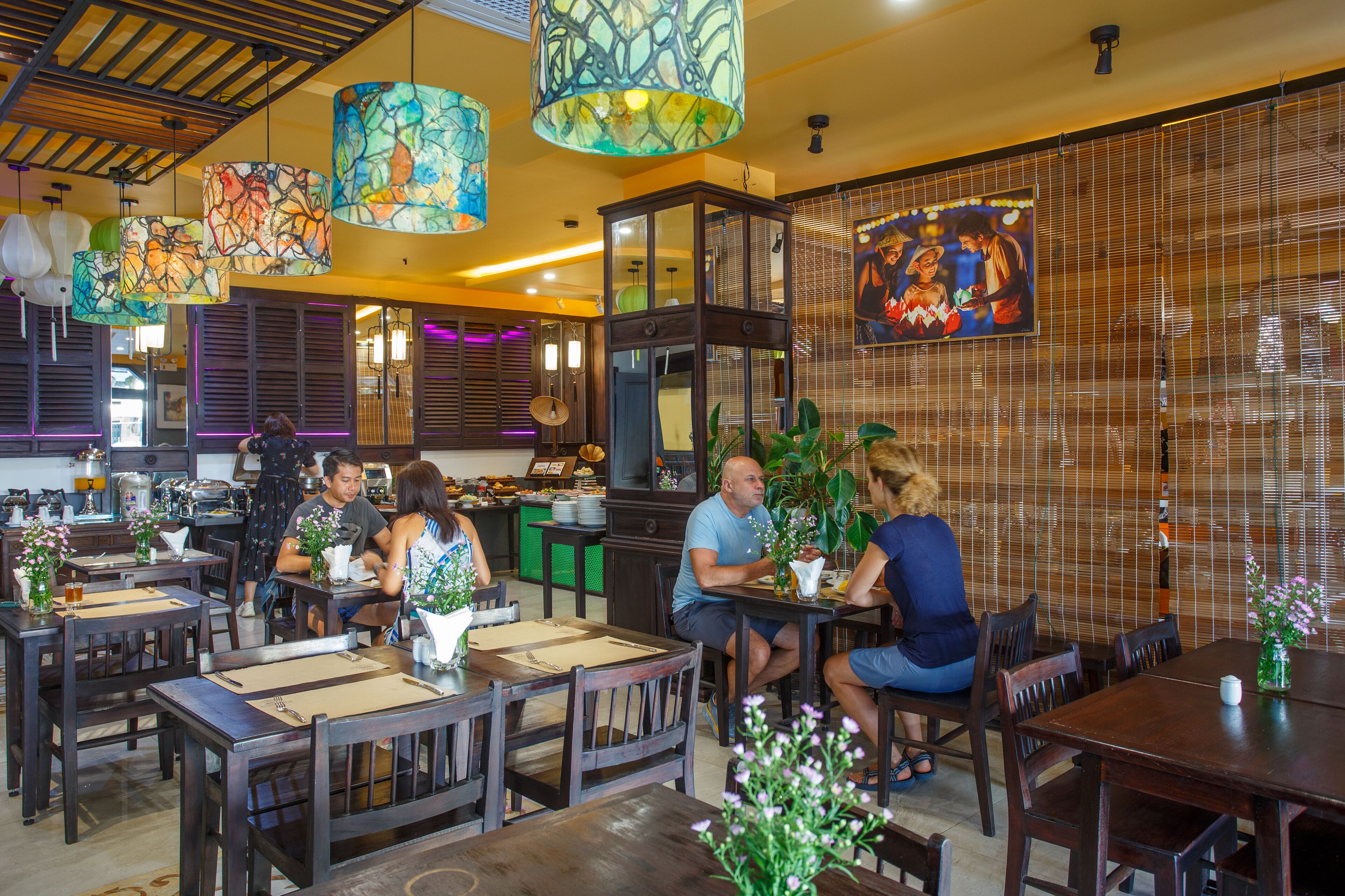 Ein Restaurant mit Holztischen und -stühlen, bunten Hängeleuchten und einem großen, gerahmten Bild an der Wand.