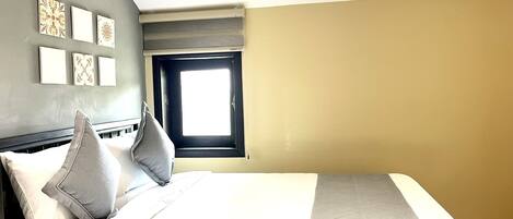 A hotel room with a bed, pillows, a window, and wall art.