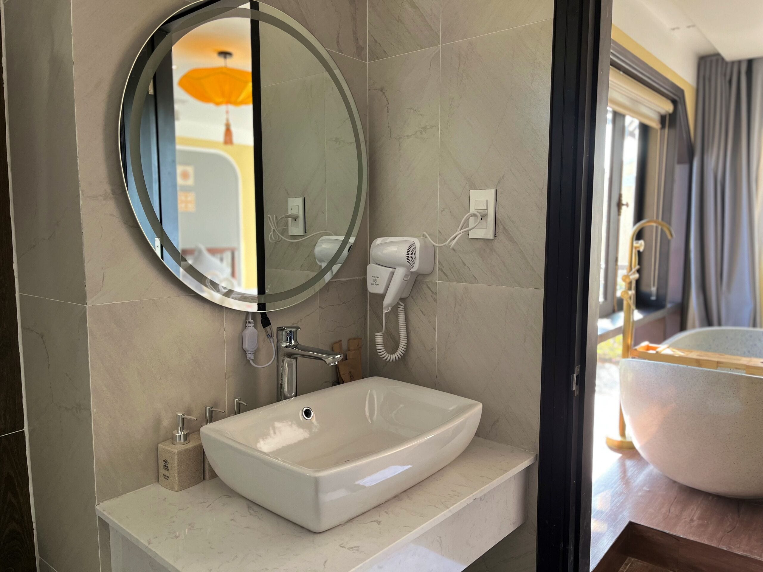 A modern bathroom with a large oval mirror, a rectangular sink, a hairdryer, and a bathtub visible through an open door.
