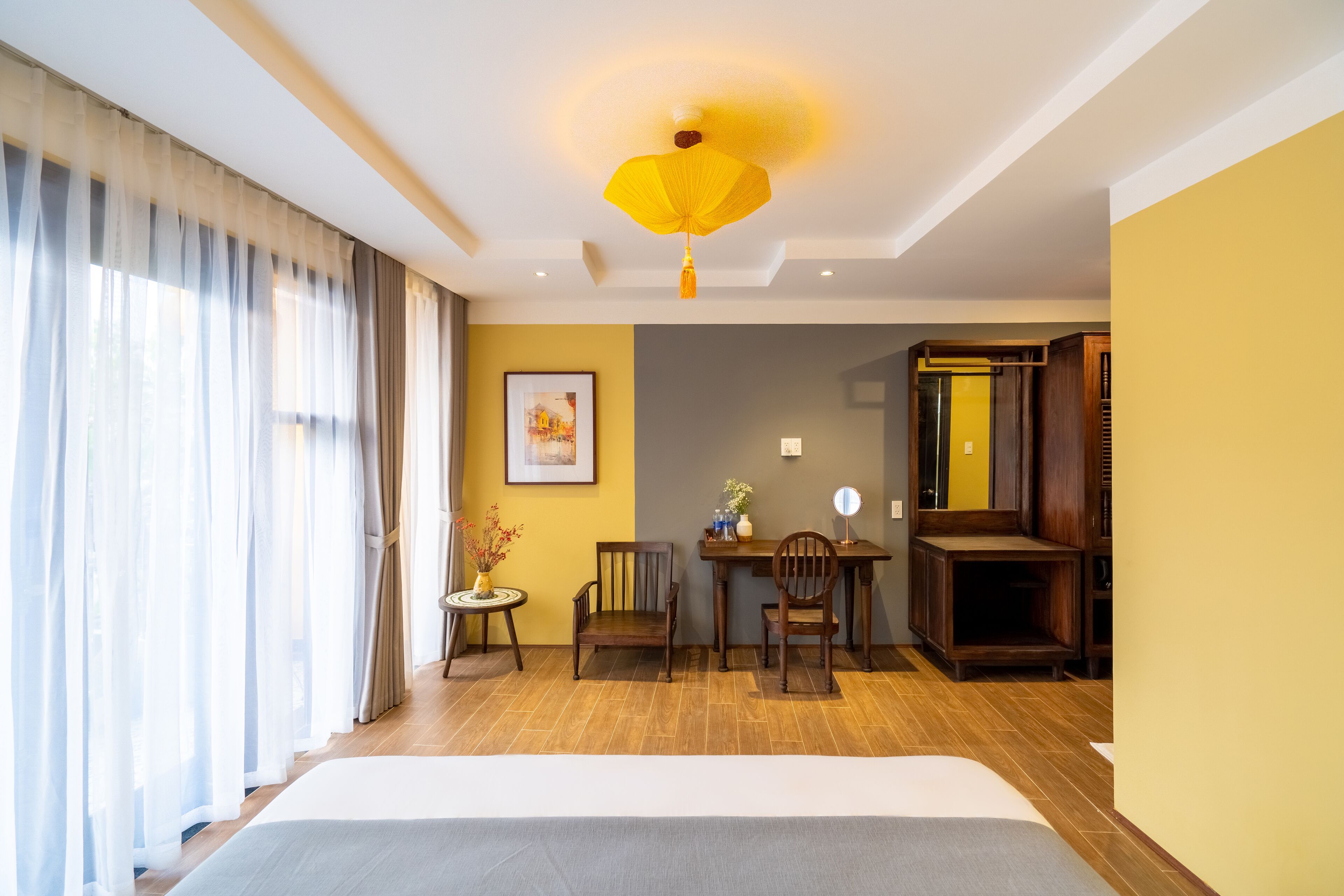 A hotel room with a bed, a desk with chairs, a small table with a vase, and a large mirror.