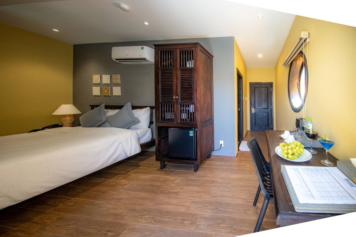 A hotel room with two beds, a wooden wardrobe, a dining table with a plate of fruit, and a bar area.