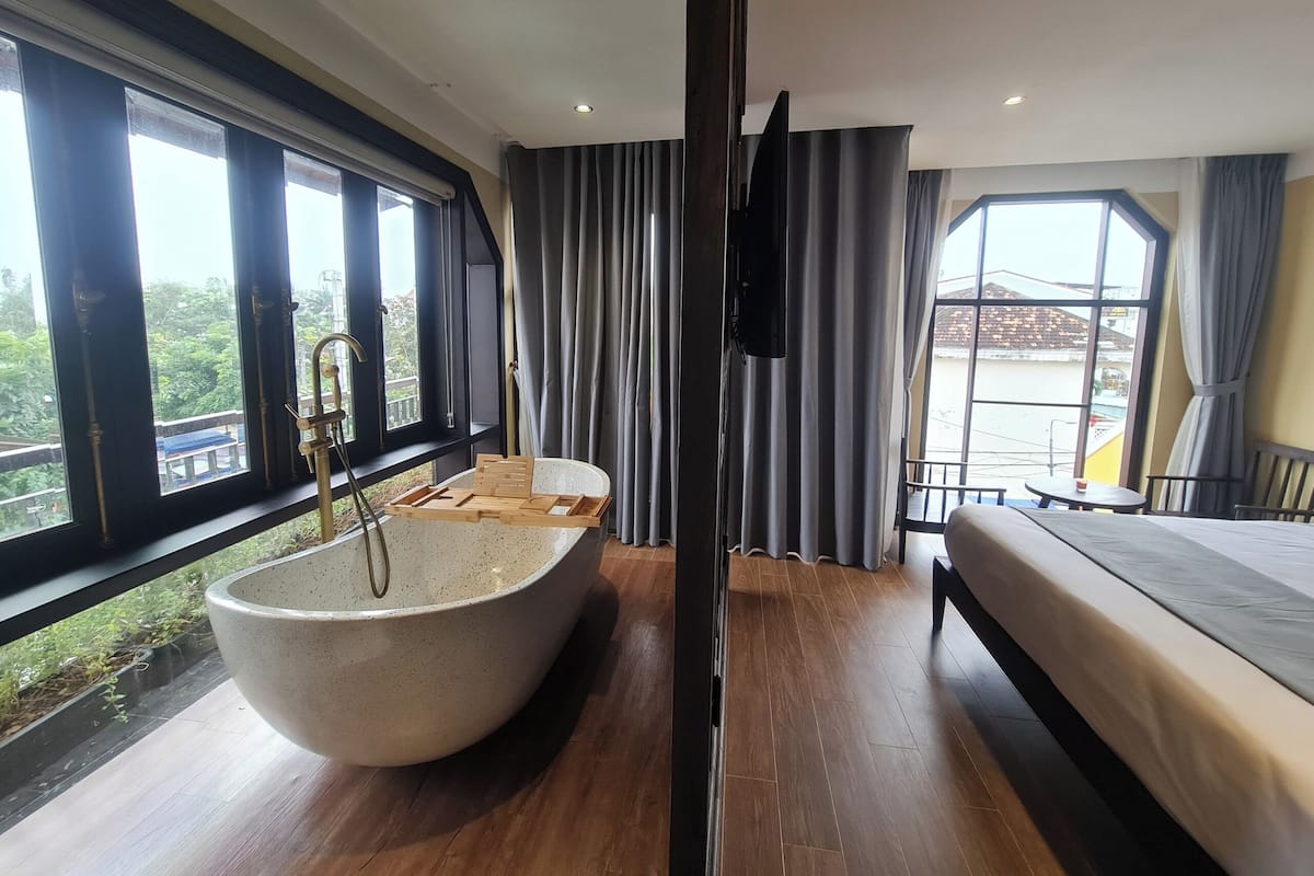 A modern bedroom with a large freestanding bathtub, a bed, and a balcony with a view of greenery.