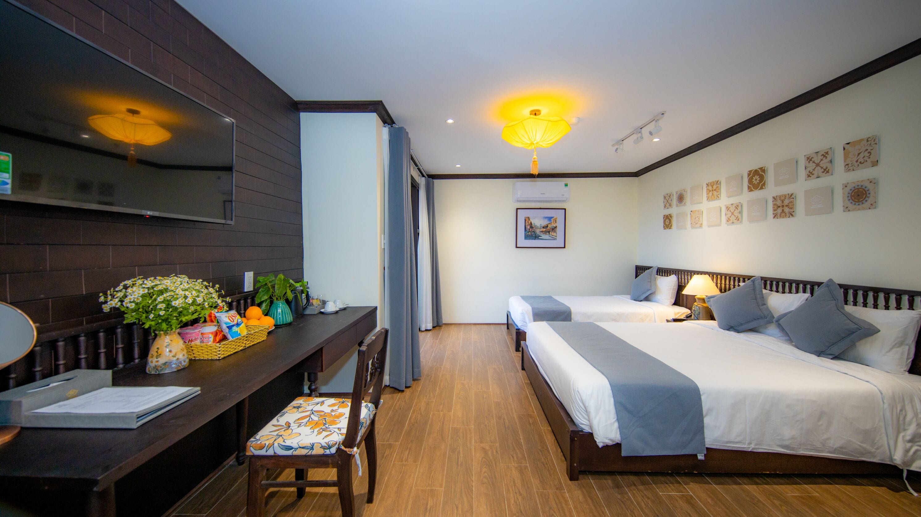 Habitación de hotel con dos camas, un escritorio con silla, una mesita con un florero y un cuadro con marcos.