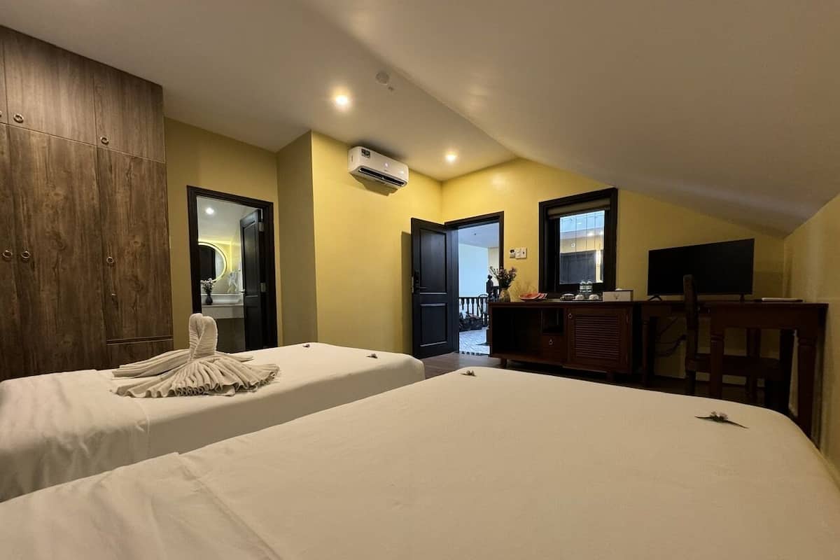 A hotel room with two beds, a wooden wardrobe, a desk with a chair, and an air conditioning unit.