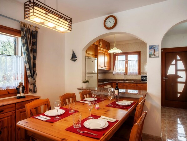 A dining area with a wooden table set for four, a kitchen in the background, and a clock on the wall.