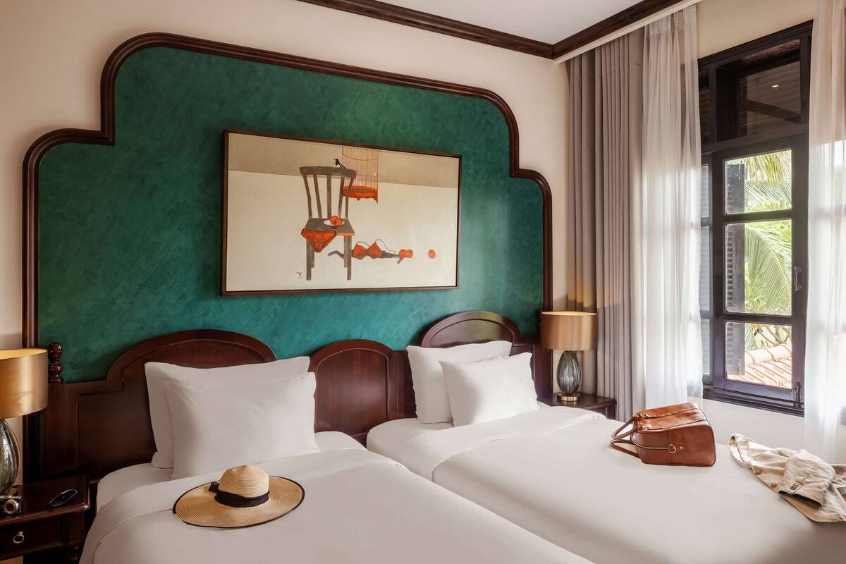 A hotel room with two beds, a large painting on the wall, a window with curtains, and a nightstand with a lamp.
