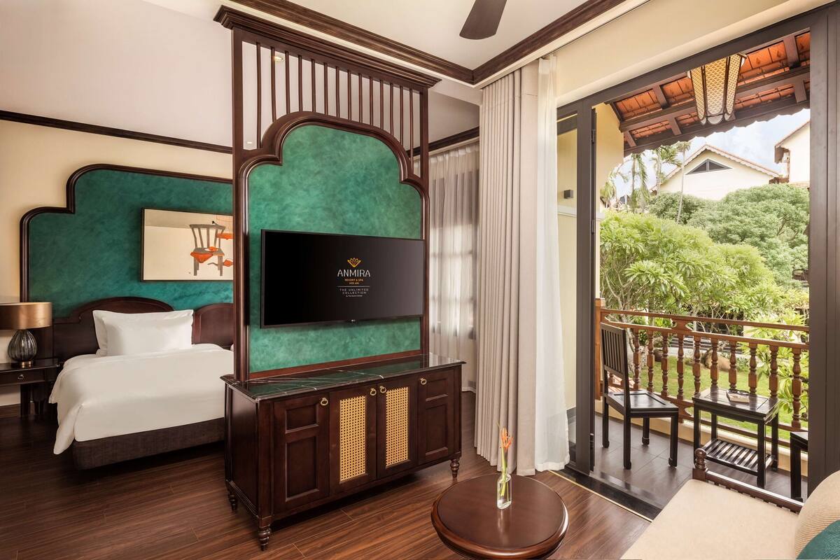 A hotel room with a bed, a TV, a wooden cabinet, a round table, and a balcony with a view of greenery.