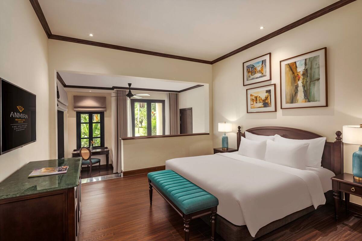 A hotel room with a large bed, a bench, a desk with a chair, a TV, and framed artwork on the wall.