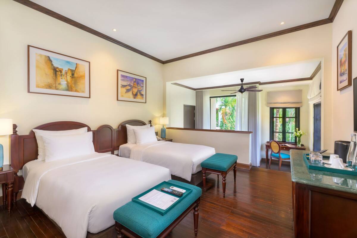 A hotel room with two beds, a desk, a chair, a window with a view, and artwork on the walls.