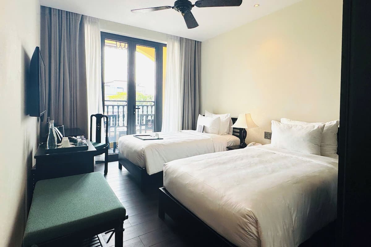 A hotel room with two beds, a desk, a chair, a ceiling fan, and a balcony with curtains.