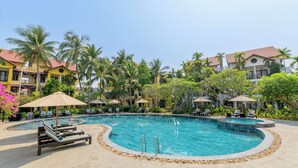Área de piscina de resort com espreguiçadeiras, guarda-sóis e uma piscina azul cristalina, cercada por palmeiras e edifícios do resort.