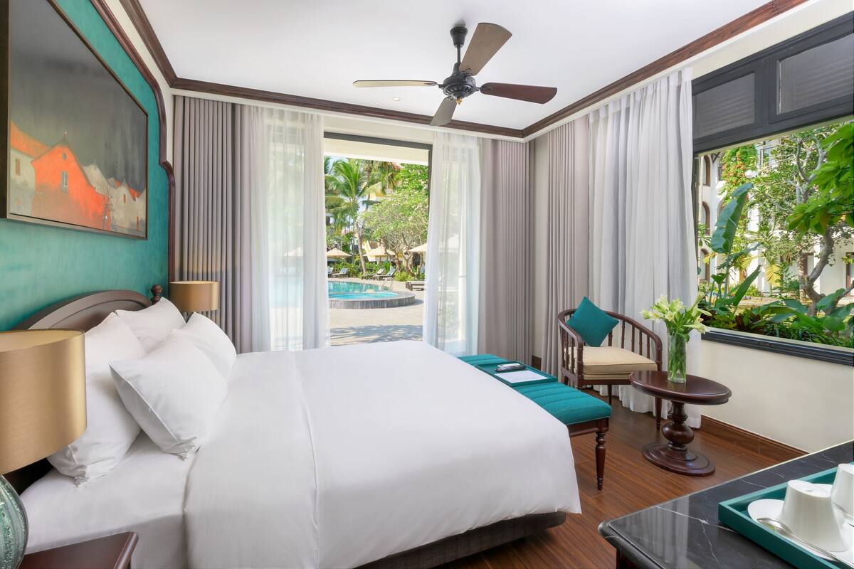 A hotel room with a large bed, a ceiling fan, a view of a pool and greenery outside.