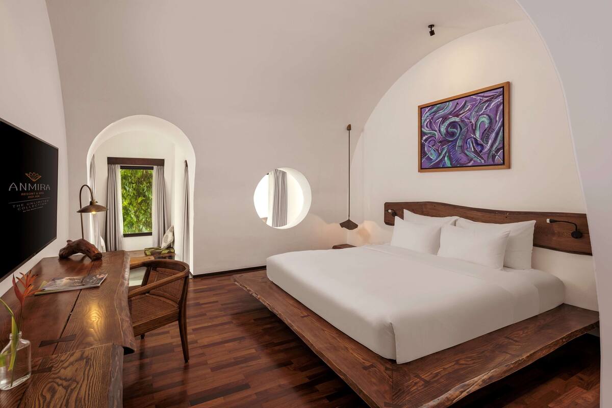 A modern hotel room with a wooden bed, a desk with a flat-screen TV, a round mirror, and a large abstract painting on the wall.