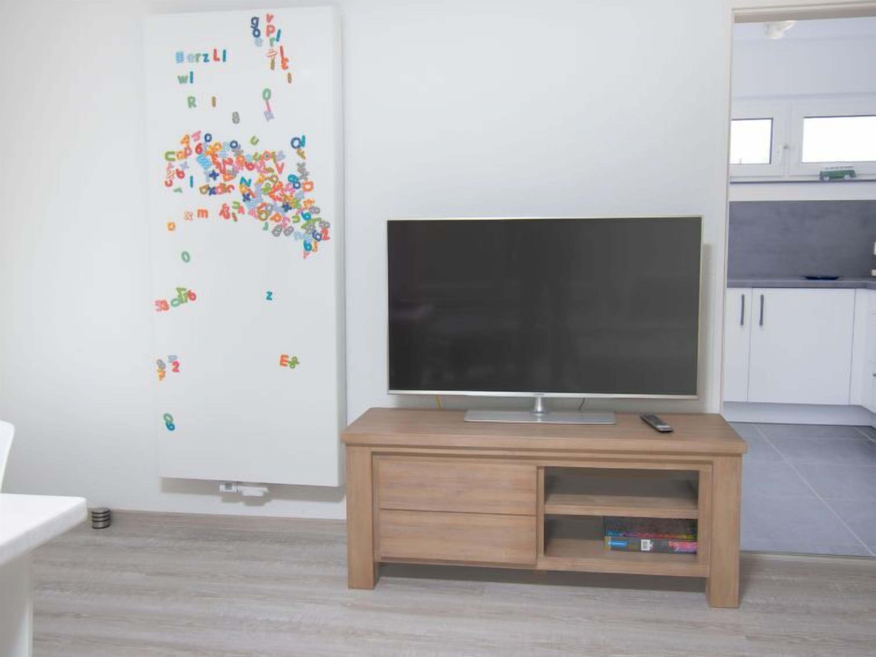 A room with a flat-screen TV on a wooden stand, a whiteboard with colorful letters, and a kitchen area with cabinets and a microwave.