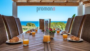 Una mesa lista para el desayuno con cruasanes, zumo de naranja y un florero, con vista al mar.