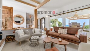 Amplia sala de estar con un sofá marrón, dos sillones, una mesa de centro y un otoman estampado. Se observa un techo con estructura de madera y un ventanal con vistas al mar.