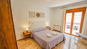 Une chambre à coucher avec une couvre-lit à rayures, des tables de chevet en bois, un radiateur et une vue sur l’extérieur, visible à travers une porte vitrée.