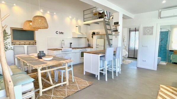 Une cuisine moderne avec des meubles blancs, un coin repas doté d’une table et de chaises en bois, et une chambre à l’étage avec un lit superposé.