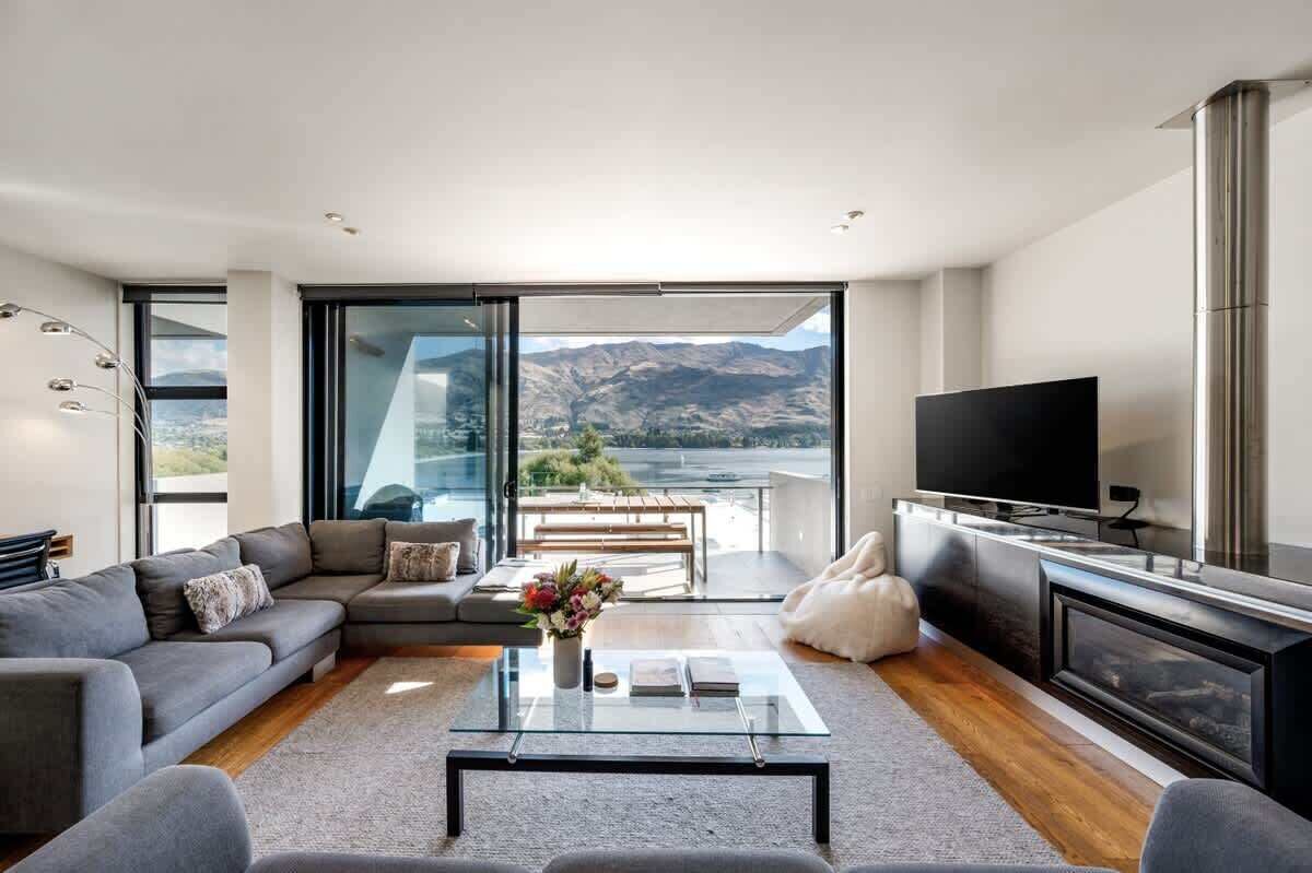 Una sala moderna con sofá, mesa de centro y televisor, un balcón con vistas a montañas y una chimenea.