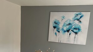 Uma área de refeições com mesa de madeira, quatro cadeiras, um vaso com plantas secas e um quadro de flores azuis na parede.