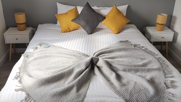 Uma cama com colcha listrada em cinza e branco, duas mesas de cabeceira com luminárias e três almofadas em amarelo e cinza.