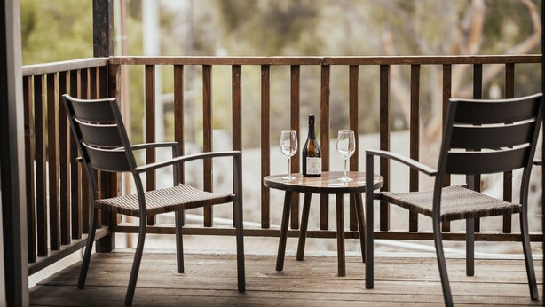 Een houten terras met twee stoelen en een kleine tafel, elk met een wijnglas en fles.