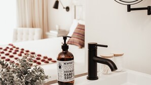 Een badkamer met een witte wastafel, een zwarte kraan en een fles handzeep. Er hangt een spiegel aan de muur en er staat een plant op de rand van de wastafel.