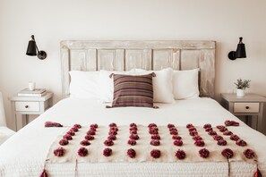 Een net opgemaakt bed met een gestreept kussen, een zachte witte deken en decoratieve pompons. Twee nachtkastjes met lampen en een kleine plant.