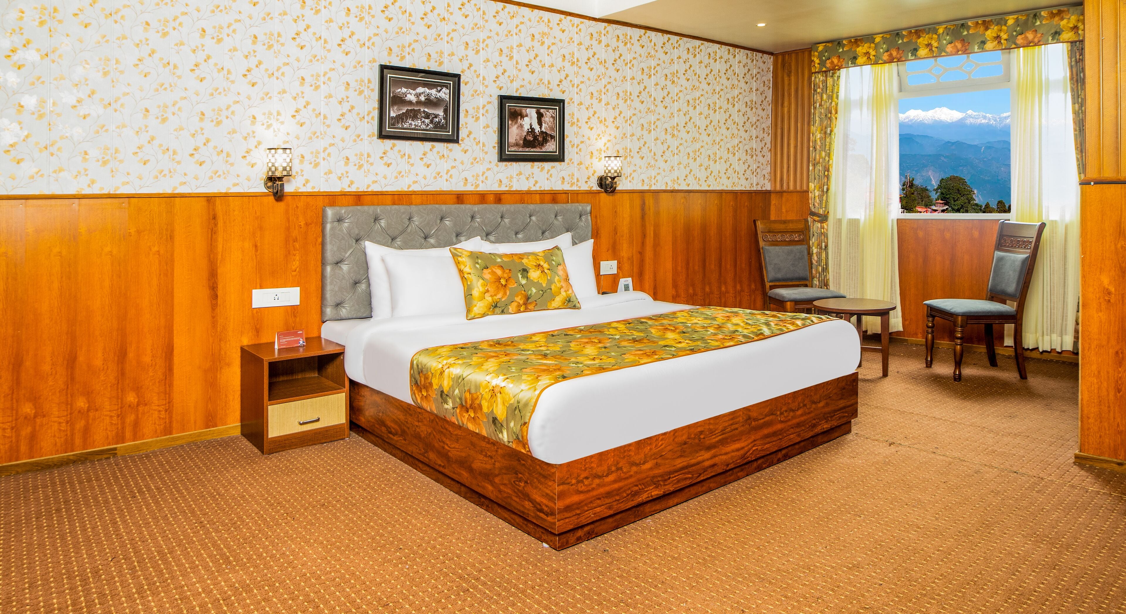 A hotel room with a large bed, a wooden headboard, a nightstand, a chair, a small table, and a view of mountains through the window.