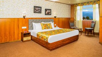 A hotel room with a large bed, a wooden headboard, a nightstand, a chair, a small table, and a view of mountains through the window.