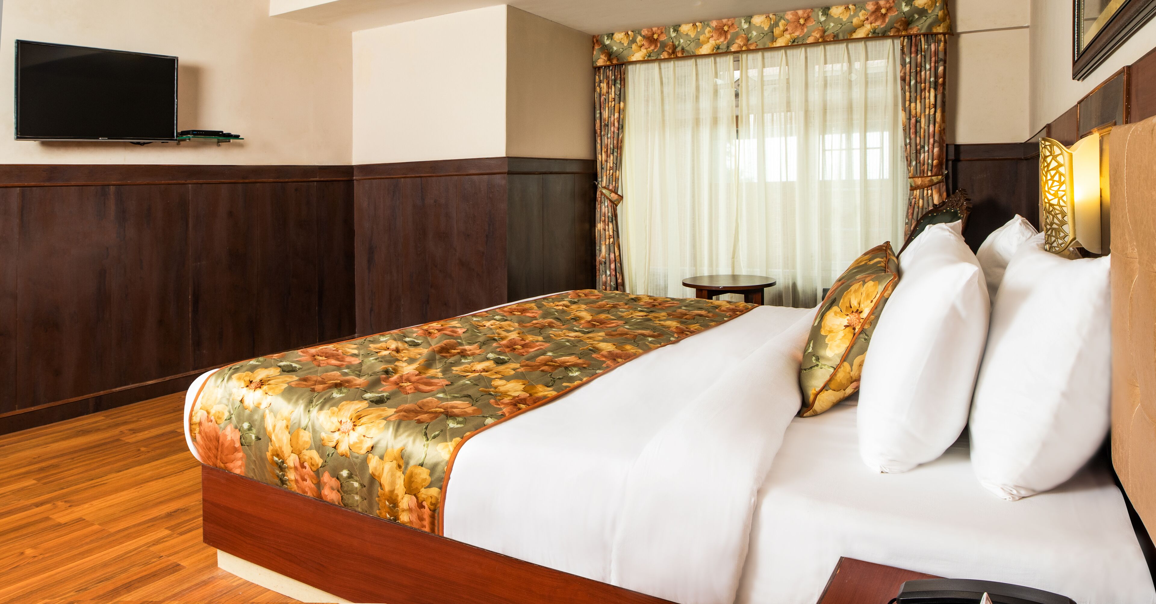 A hotel room with a wooden floor, a bed with floral bedding, a television on the wall, and a window with curtains.