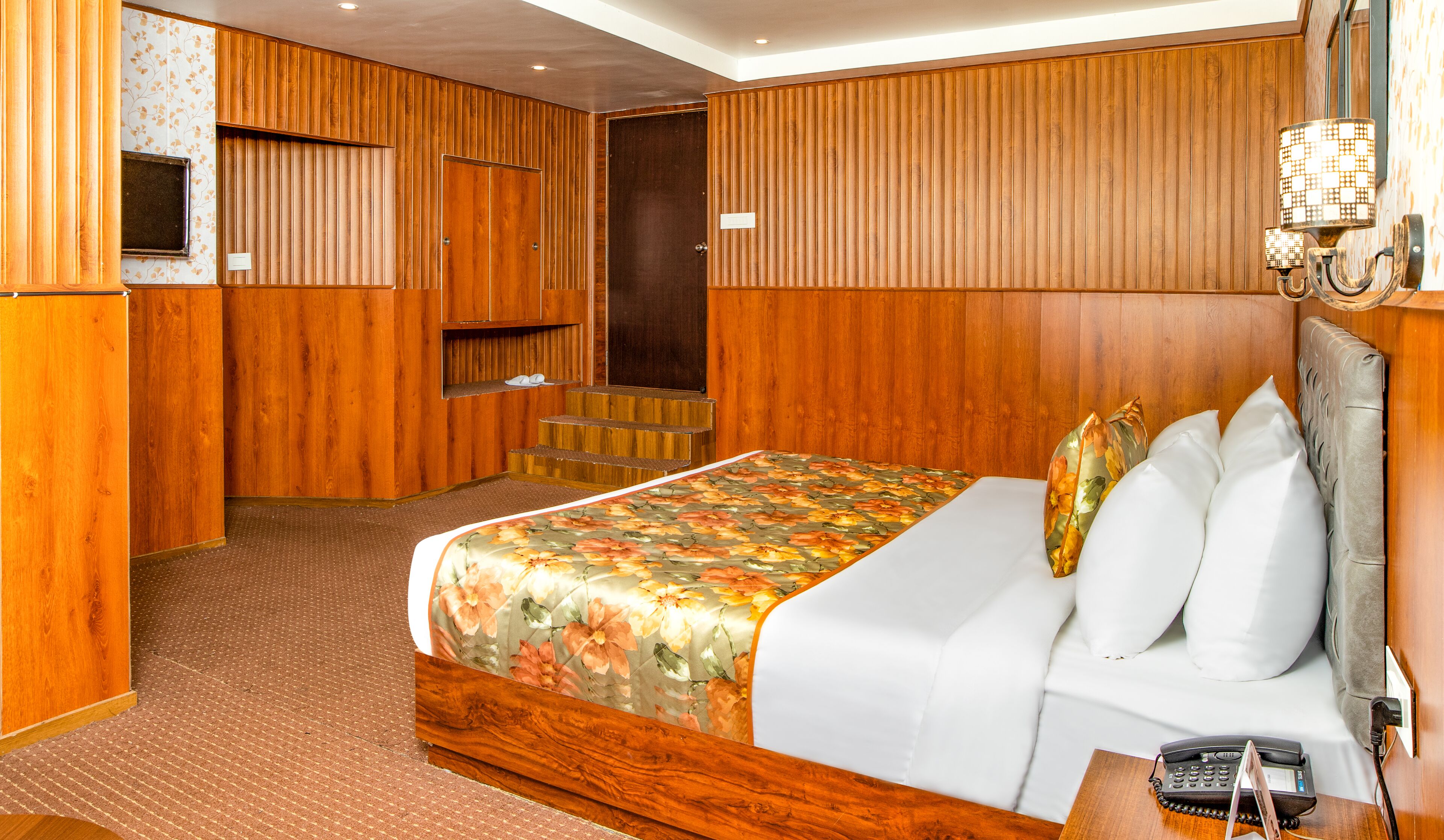 A hotel room with a wooden panel wall, a bed with floral bedding, a bedside telephone, and a shoe rack.