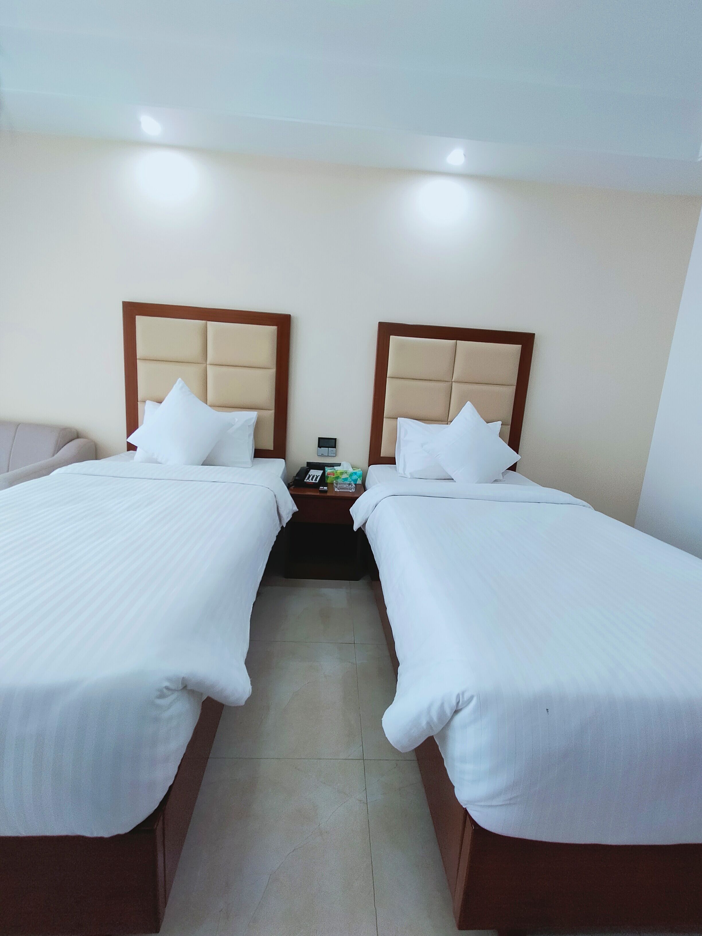 A hotel room with two single beds, each with a wooden headboard and white bedding. There is a nightstand between the beds with a phone and a small item on it.