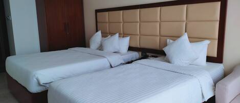 A hotel room with two single beds, a wooden wardrobe, and a large padded headboard.