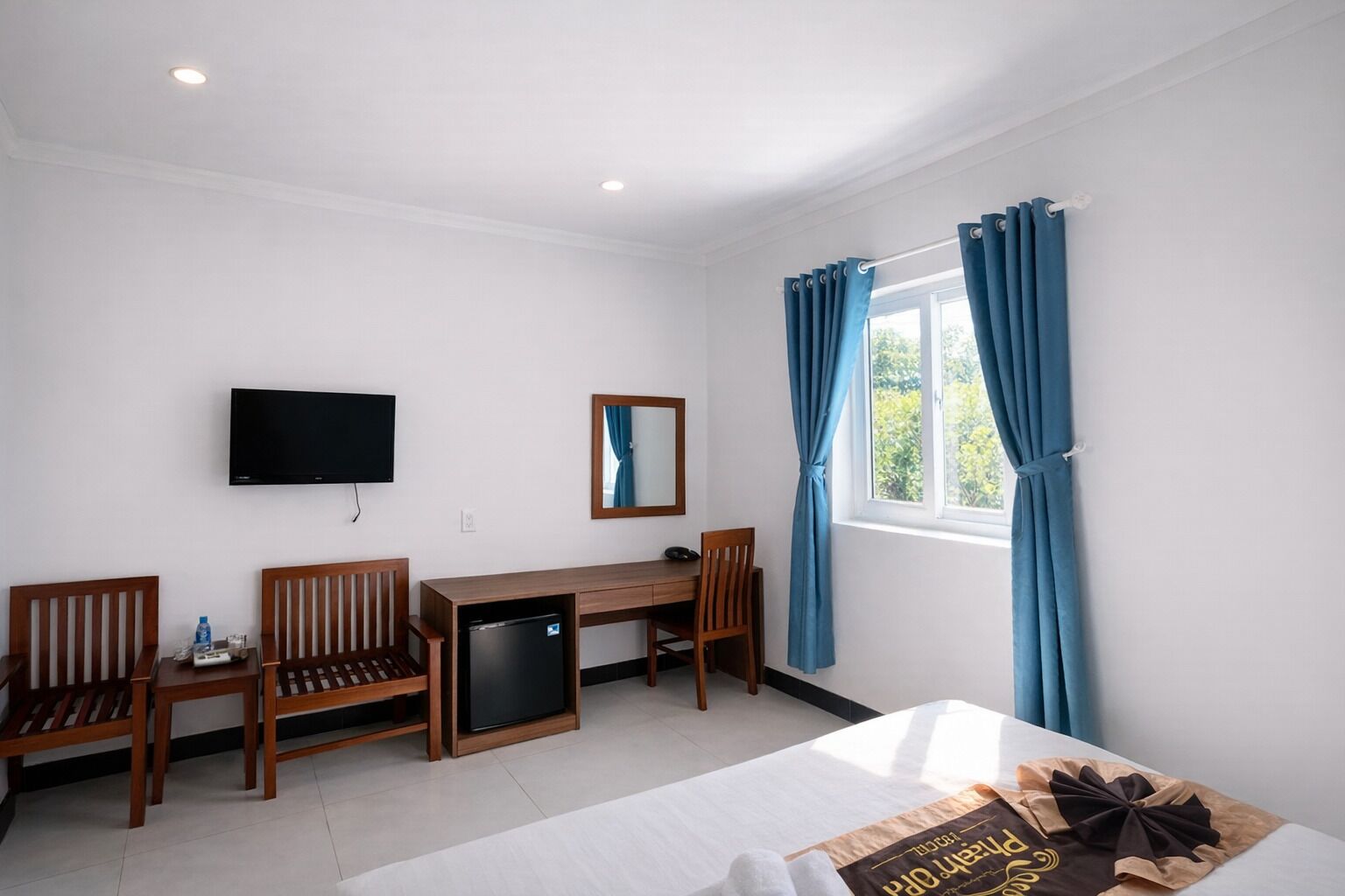 A hotel room with a bed, a desk, a chair, a TV, a small fridge, and a window with curtains.