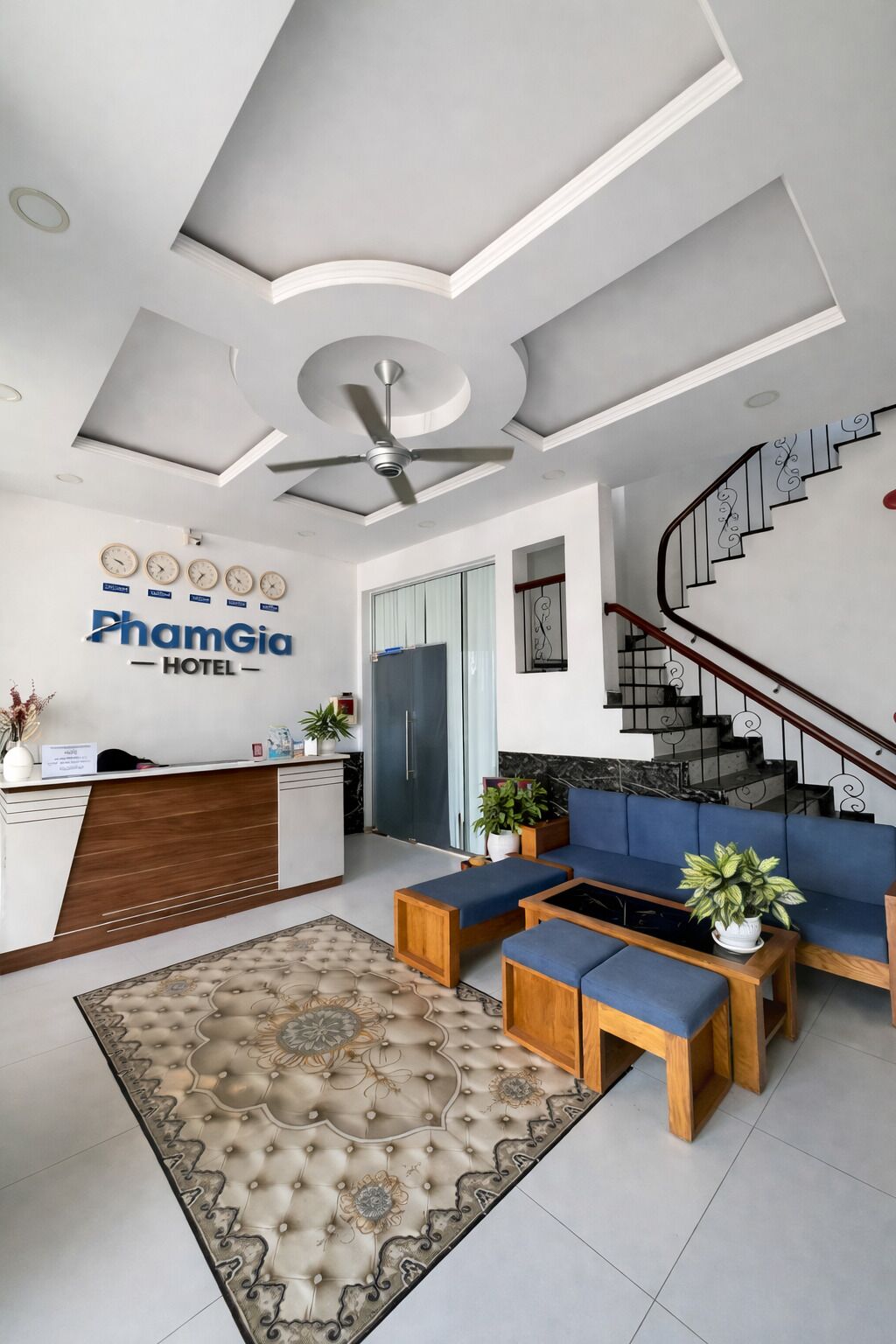 Un vestíbulo de hotel con mostrador de recepción, una zona de estar con sofás y una mesa de centro, una escalera y un ventilador de techo.