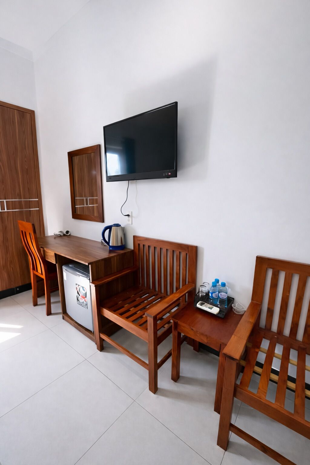 Una habitación con un escritorio de madera, un televisor montado en la pared, un refrigerador pequeño y dos sillas de madera.