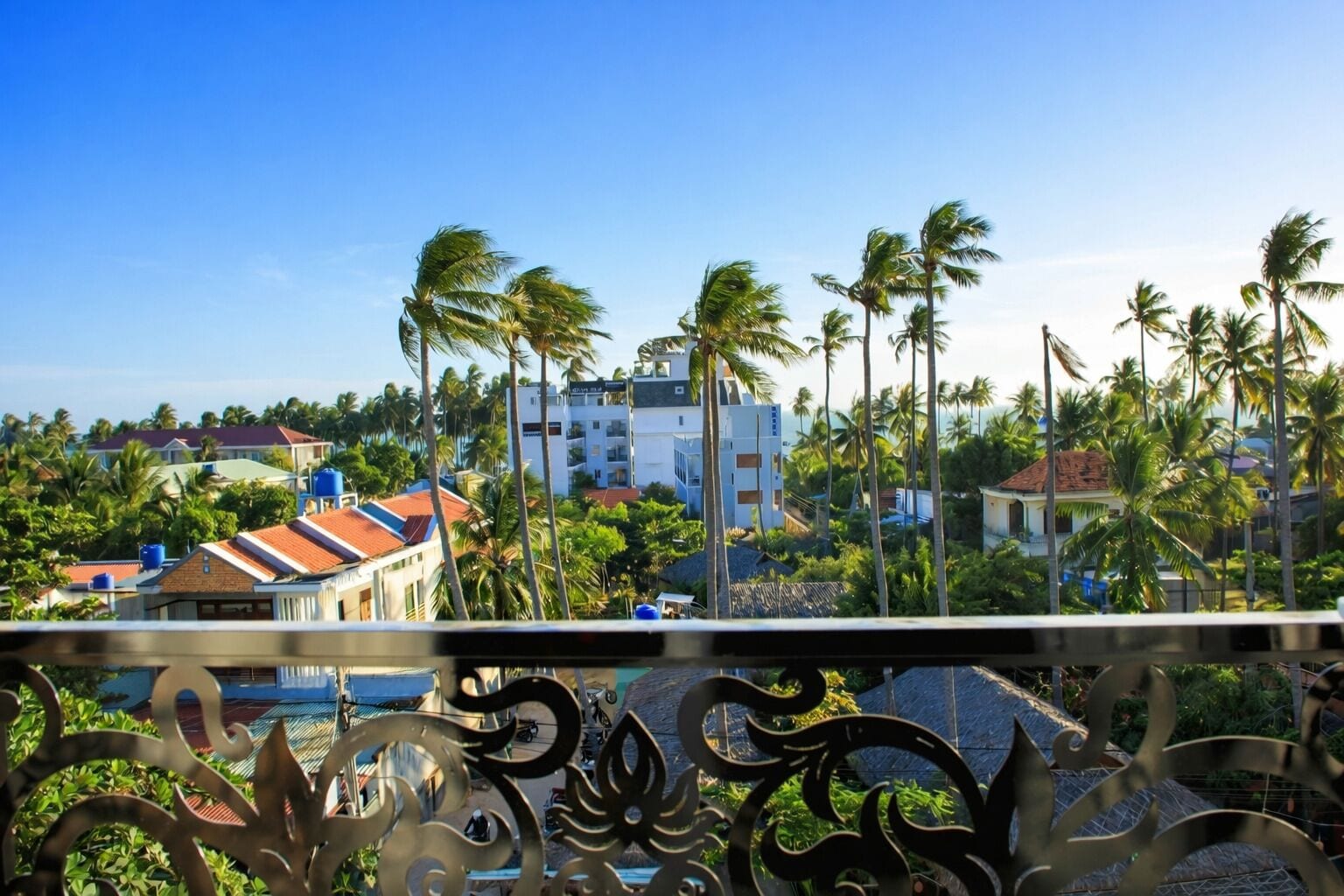 Vista costera con palmeras, un edificio de techo rojo y un balcón con barandillas de metal decorativo.