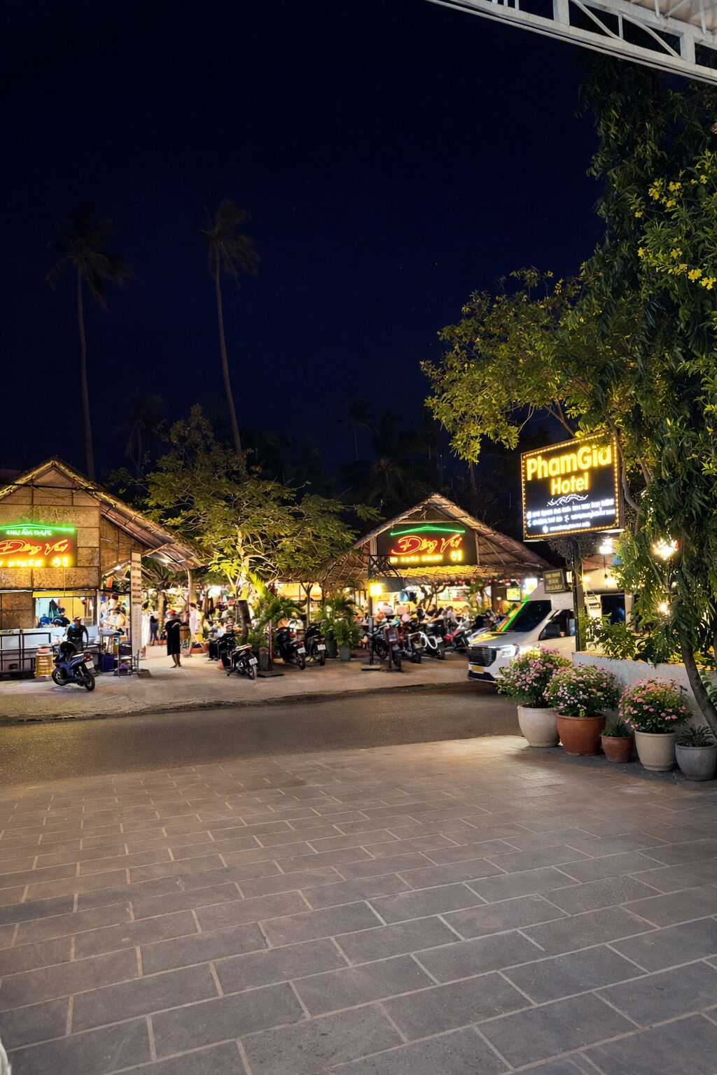 A night scene at a hotel with a sign for 'PhamGia Hotel,' a covered outdoor seating area, and several parked motorcycles.
