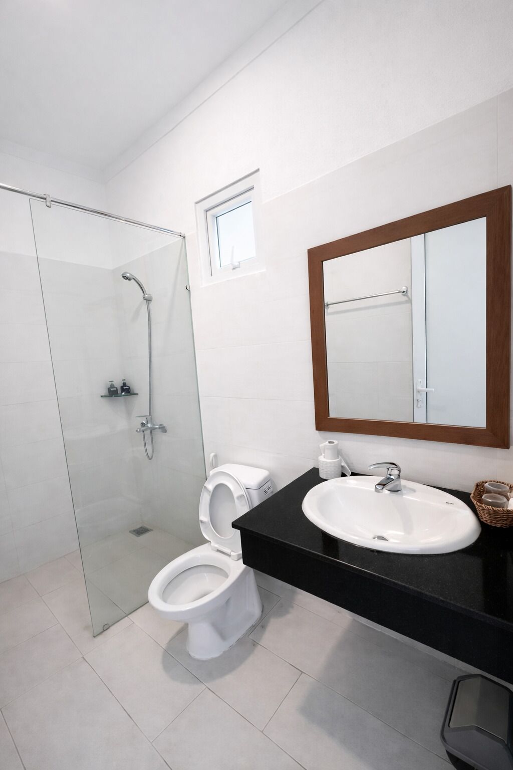 A modern bathroom with a glass shower enclosure, a toilet, a sink with a mirror, and a small window above the toilet.