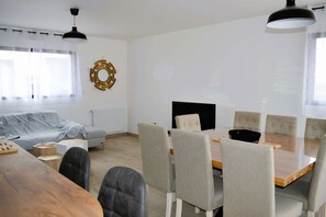 A modern living room with a sofa, a dining table with chairs, a fireplace, and a large window with curtains. - La Mer Veille, new house with garden (Courseulles-sur-Mer)