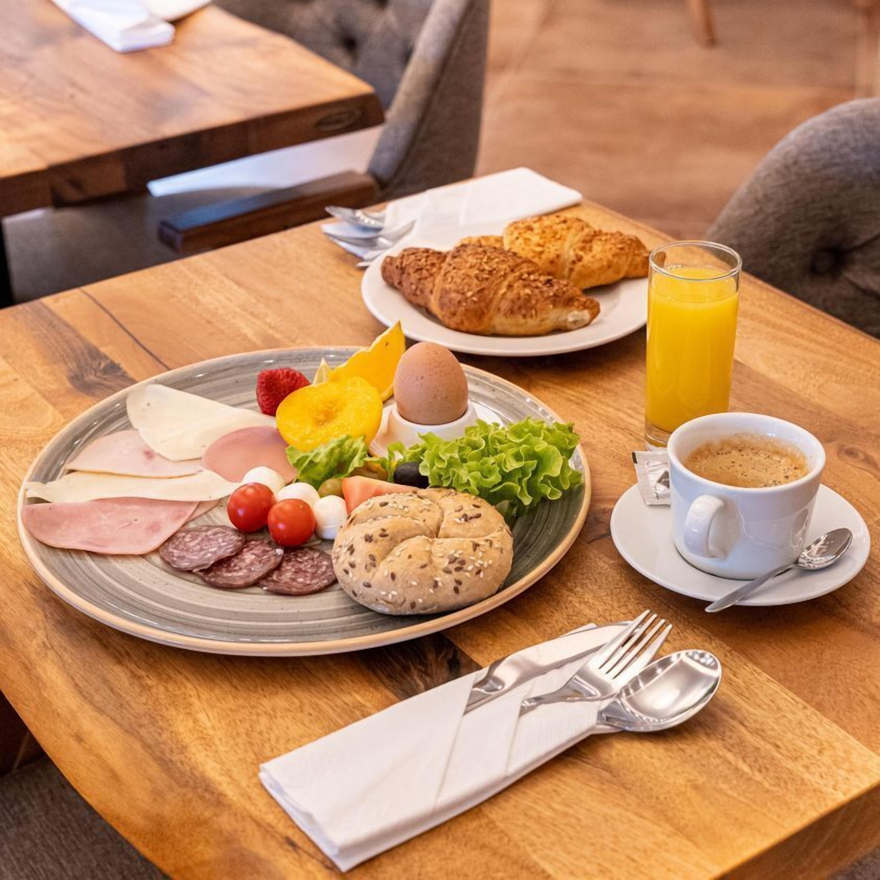 Een ontbijtsetting met een bord eten, croissants, sinaasappelsap en een kop koffie op een houten tafel.