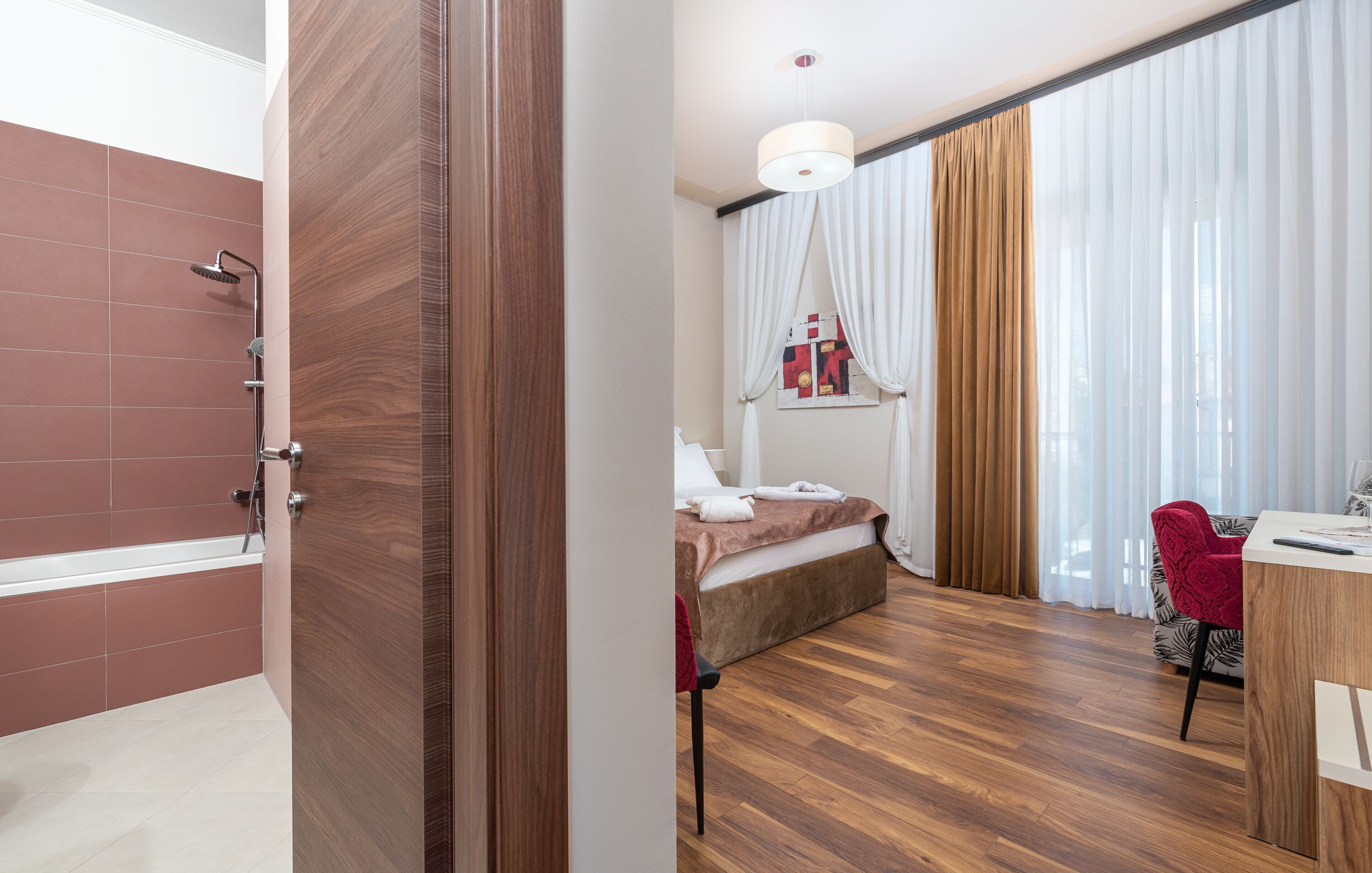 A hotel room with a wooden floor, a bed, a desk with a chair, and a bathroom with a shower and a bathtub.