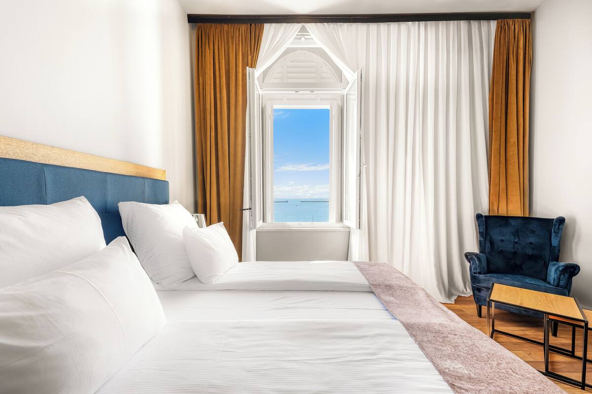 A hotel room with a large bed, a chair, and a view of the sea through an open window.