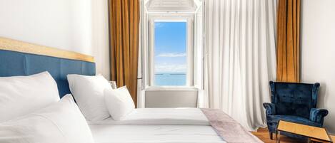 A hotel room with a large bed, a chair, and a view of the sea through an open window.