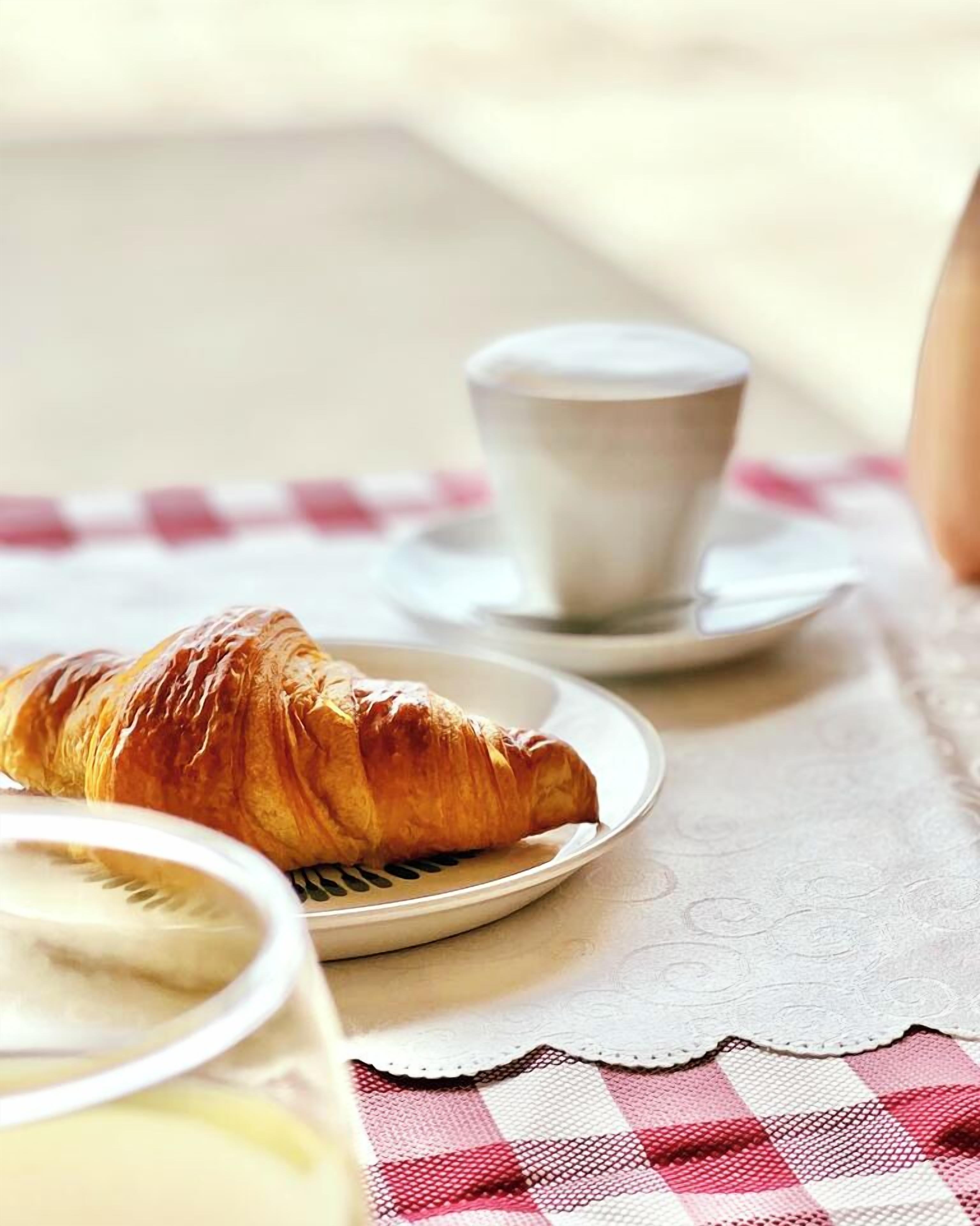 Een croissant op een bord, een kop koffie en een glas melk op een tafel met een geruit tafelkleed.