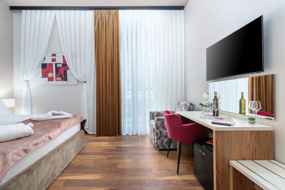 A modern hotel room with a bed, a desk with a chair, a TV, and a window with curtains.