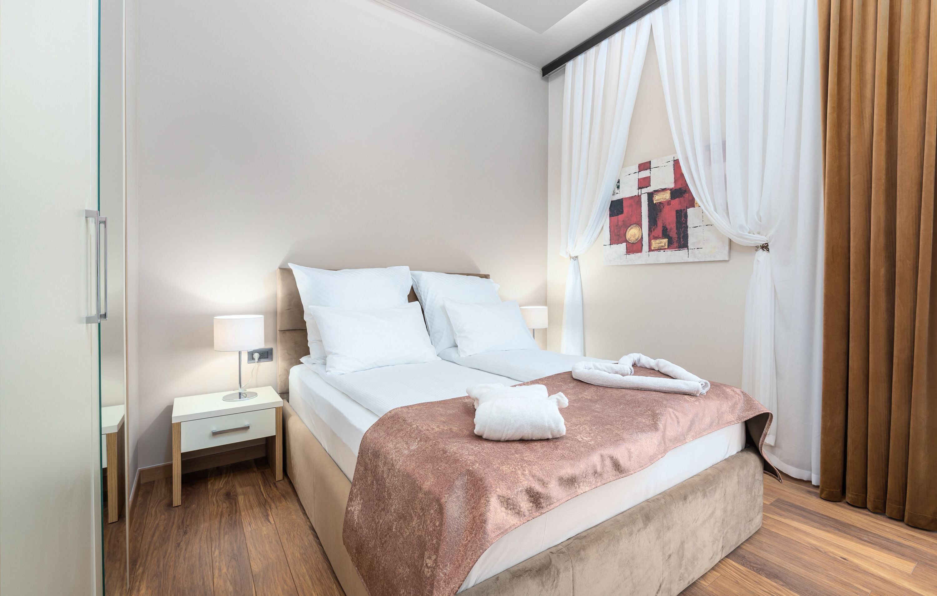 A neatly made bed with white linens and a pink blanket, a bedside table with a lamp, and a wall-mounted artwork.