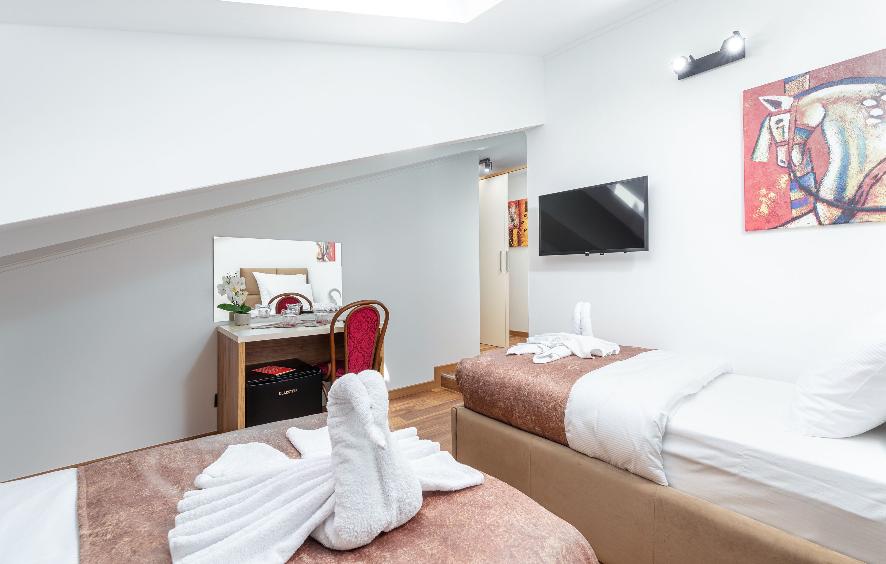 A modern hotel room with a bed, a desk with a chair, a TV, and a painting on the wall.