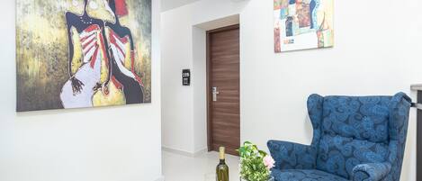 A room with a blue patterned armchair, a round wooden table with a bottle and glass, and two artworks on the walls.